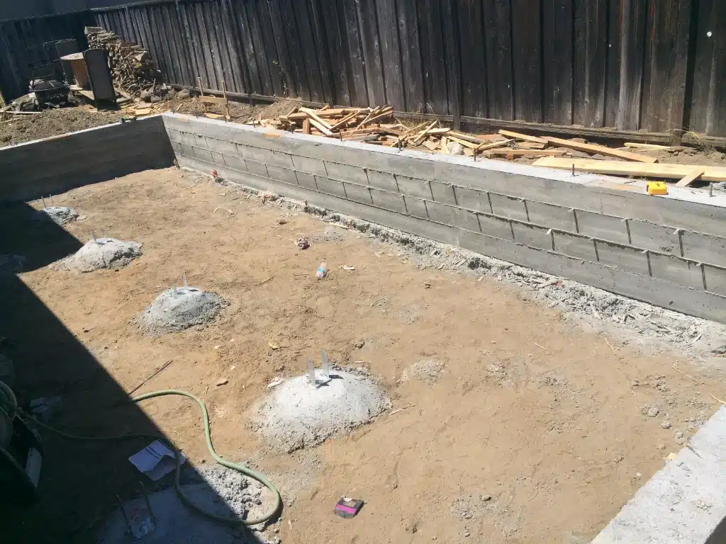 Concrete foundation piers set in a dirt area beside a new block retaining wall with scattered lumber nearby.