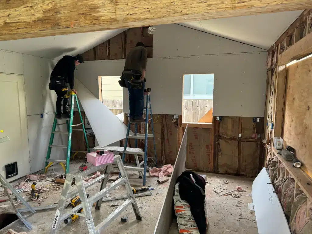 Contractors installing drywall on a residential interior wall with exposed insulation and ladders.
