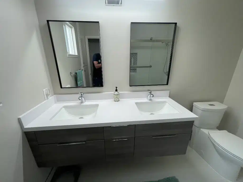 Dual-sink bathroom vanity with two mirrors, white countertop, gray cabinets, and a toilet on the right.