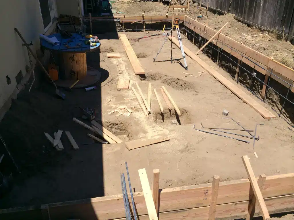 Framed foundation area with dirt excavation, scattered lumber, rebar, and a tripod level during early construction.