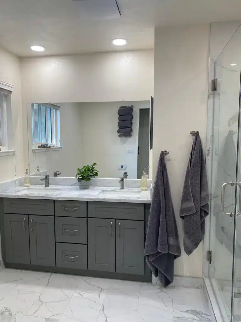 Gray double-sink vanity with marble countertop, large mirror, gray towels, and a glass shower on a white marble floor.