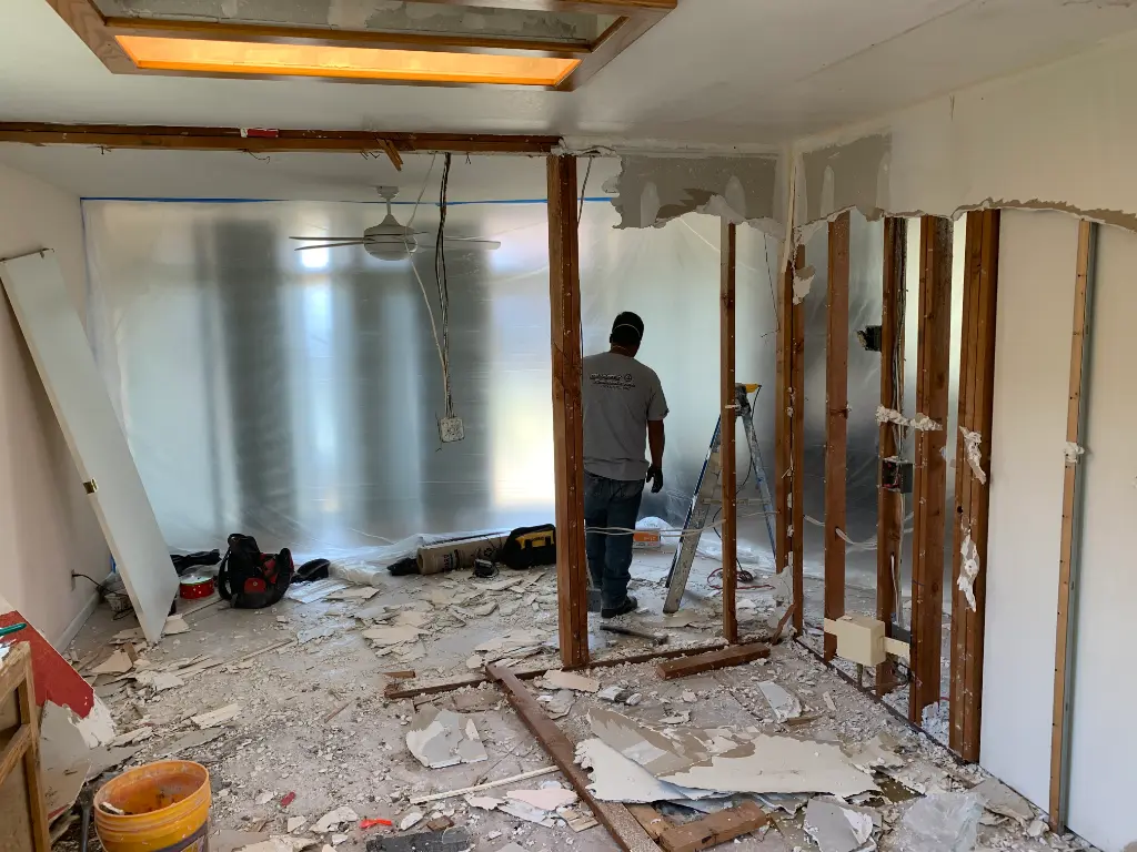Worker removing interior walls in a home under renovation, with exposed studs, debris, and protective plastic sheeting