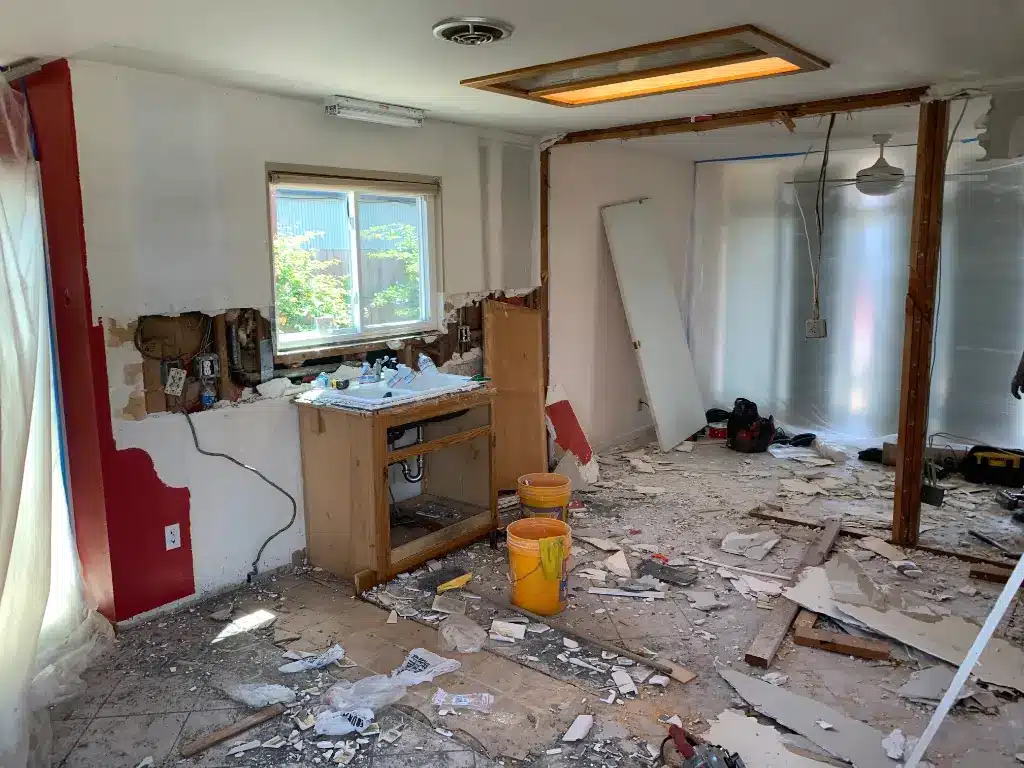 Kitchen demolition scene with torn drywall, exposed wiring, broken cabinets, debris, and tools scattered on the floor