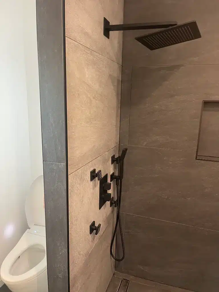 Matte black shower fixtures with rainhead and handheld sprayer beside a toilet in a modern tiled bathroom