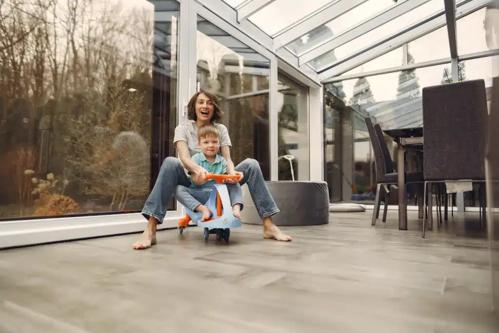 Smiling woman and young boy playing on a toy car inside a bright glass conservatory with modern furnishings.
